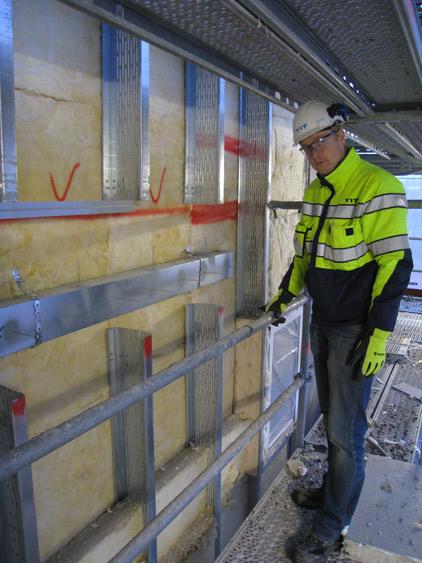 Markkinatien projektissa työnjohtajana toimiva Antti Lindroos esittelee ulkoseinärakenteen sisään asennettuja galvanoituja ilmanvaihtoputkia. Reiällisen teräsrangan väliin asennetaan vielä 200 mm lämmöneristettä ja sementtikuitulevy pintaan.