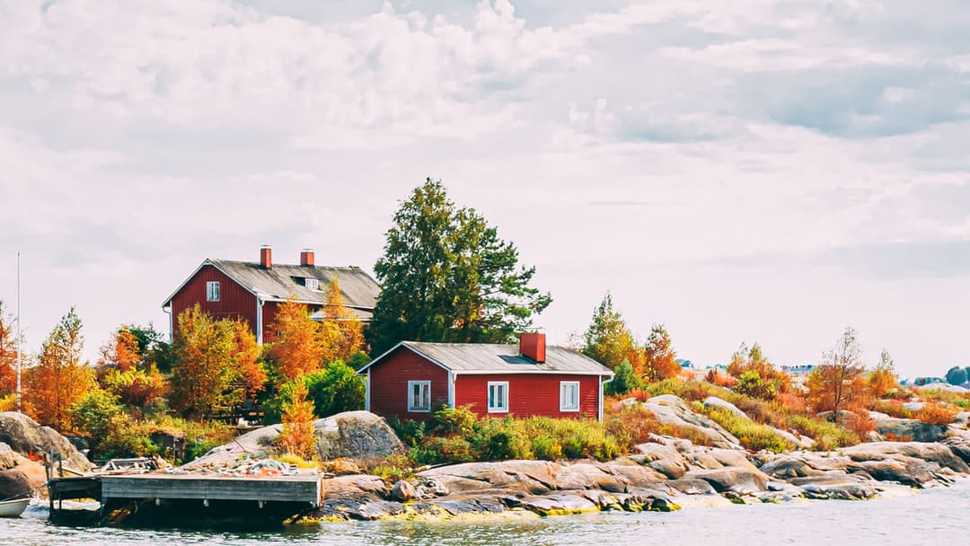 Kaksi punaista mökkiä ja laituri kauniissa saaressa jossa ruskan värittämiä puita.